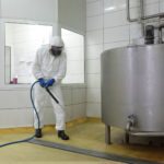 worker in white protective uniform,mask,gloves  with high pressure washer at  large industrial process tank  cleaning floor  in plant